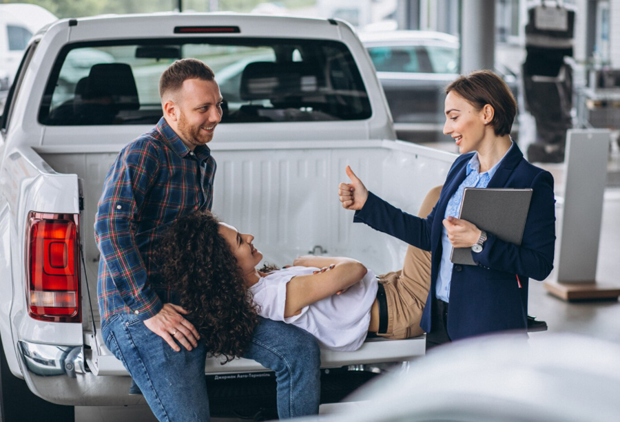 A couple seated in the bed of a pickup truck at a dealership with an agent giving a thumbs up, adding a new vehicle under Fleet Insurance Florida.