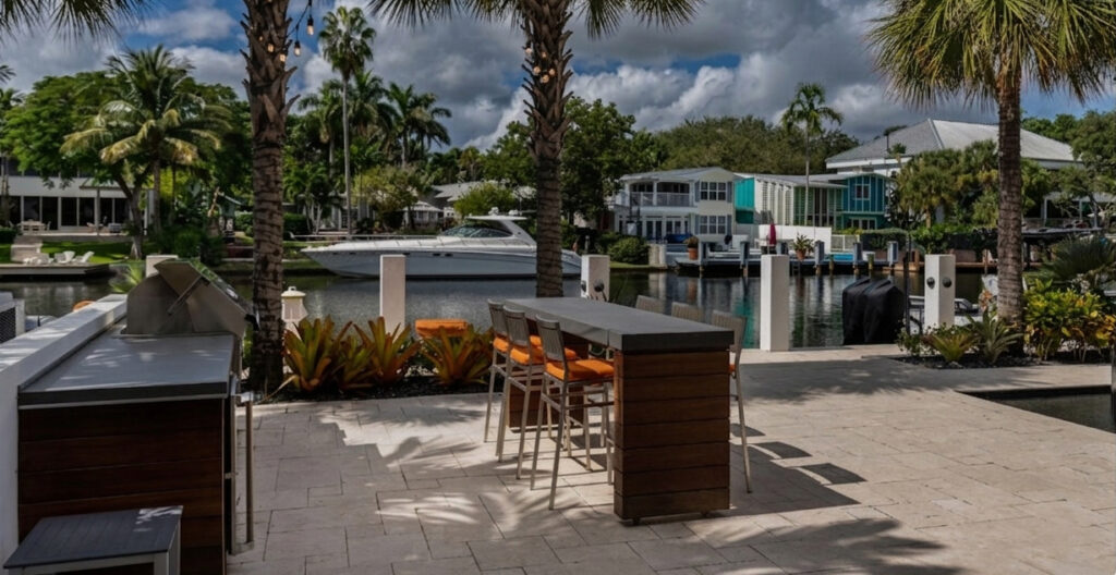 An outdoor kitchen and bar seating area beside a canal with a docked yacht and waterfront homes, the type of property General Liability Insurance protects.