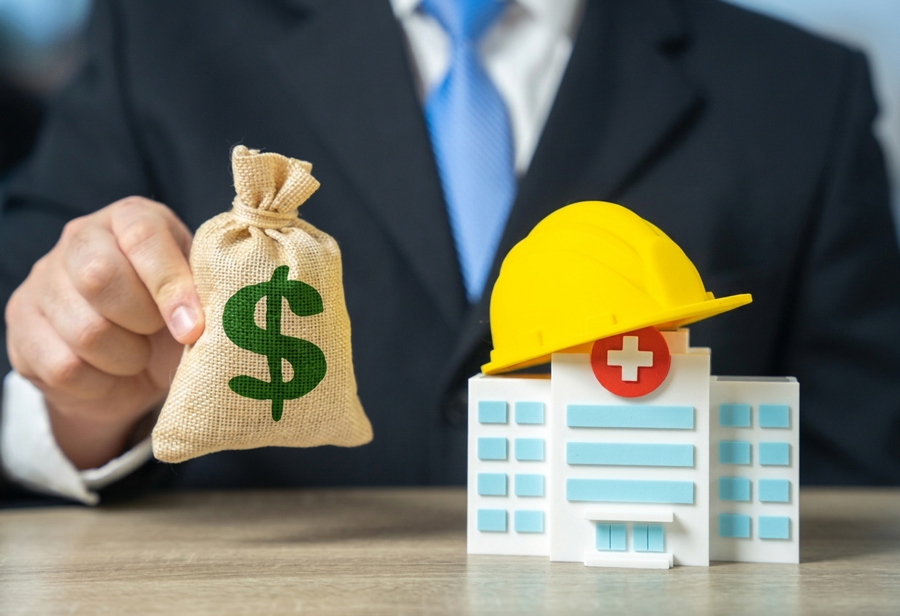 A businessman holding a money bag beside a model hospital, the medical costs 3A Allied Insurance covers with Workers Compensation Insurance Florida.