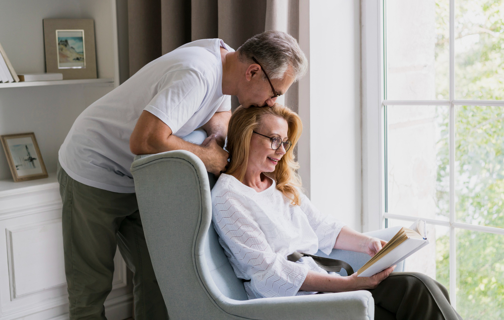 Older couple at home by a bright window, the husband leaning in to talk with his wife as she reads — representing personal insurance coverage from 3A Allied Insurance in Florida