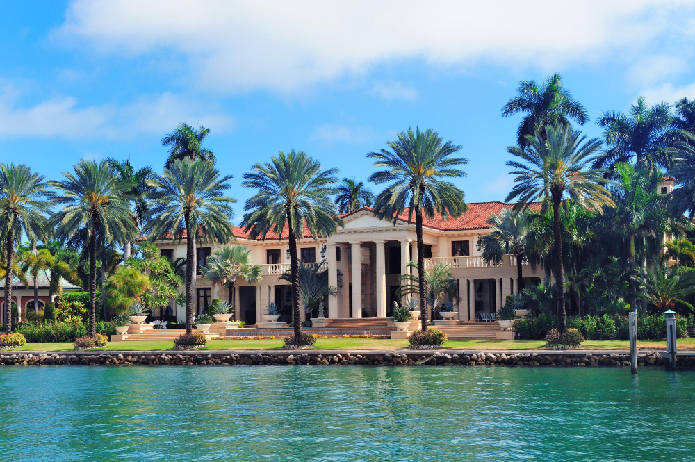 A grand columned waterfront mansion lined with palm trees sits along the water, reflecting the Real Estate Insurance Florida coverage 3A Allied provides.